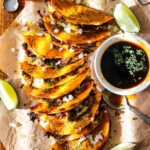 Golden crispy birria tacos on a rustic wooden board with small bowls of rich red consomm?, garnished with white onion, cilantro, and lime wedges