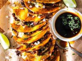 Golden crispy birria tacos on a rustic wooden board with small bowls of rich red consomm?, garnished with white onion, cilantro, and lime wedges
