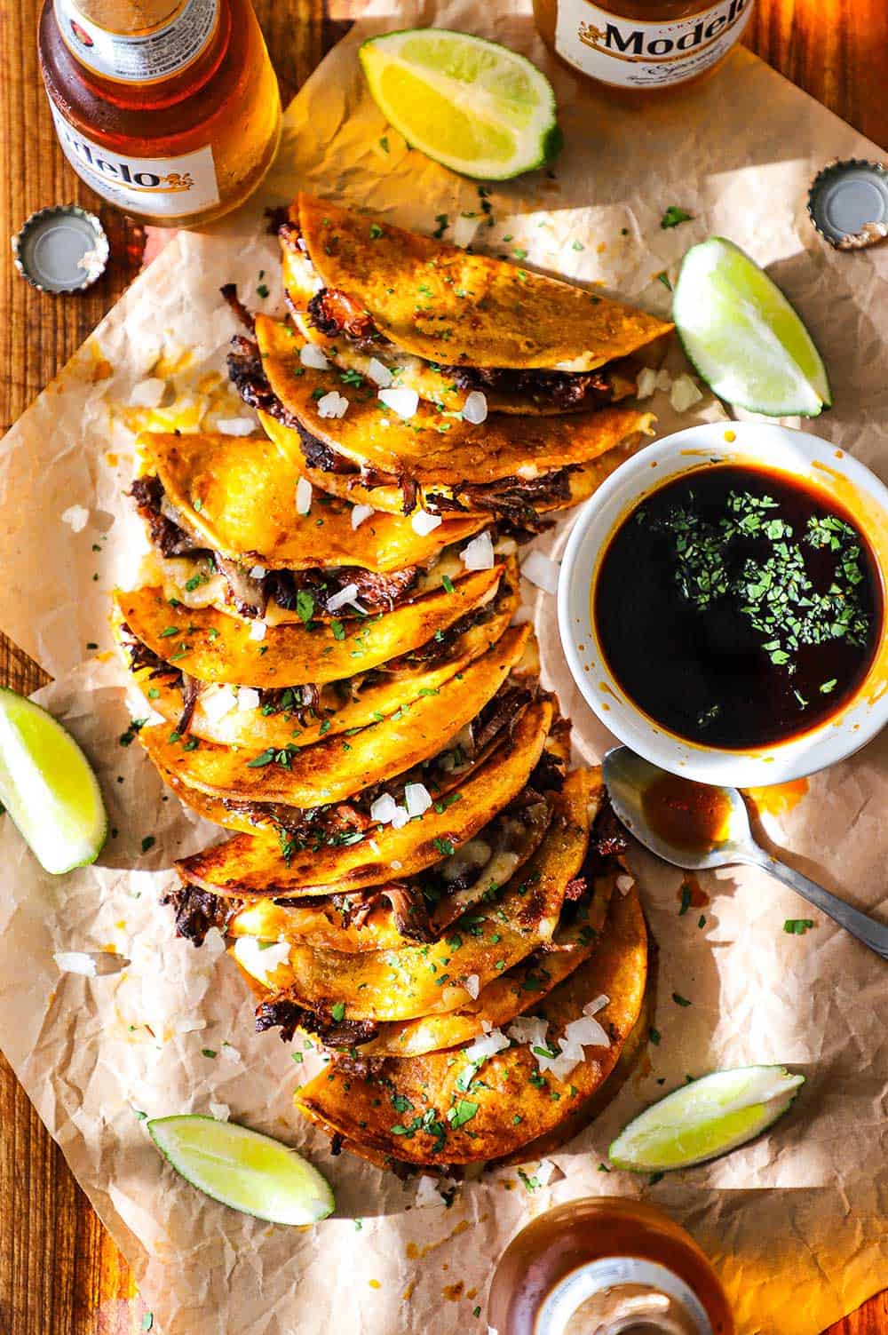 Golden crispy birria tacos on a rustic wooden board with small bowls of rich red consomm?, garnished with white onion, cilantro, and lime wedges