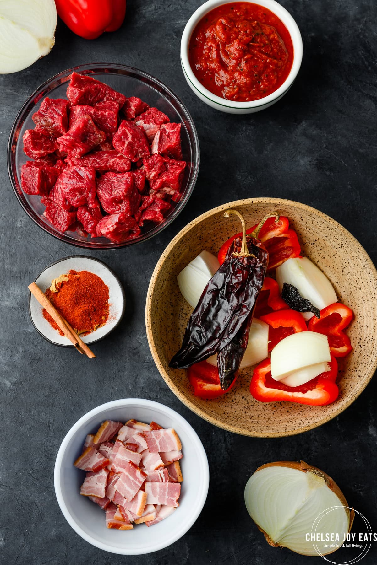 Dried guajillo, ancho, and chipotle chiles arranged on a wooden cutting board next to chunks of raw chuck roast and quartered roma tomatoes