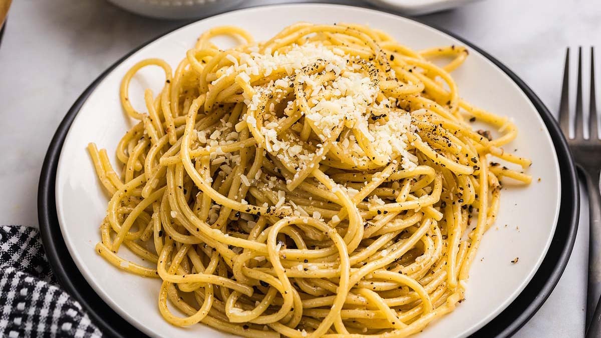 Hands grating fresh pecorino romano cheese over a bowl with freshly ground black pepper and pasta cooking in background