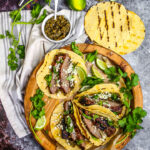 Beautifully plated carne asada tacos with charred beef, fresh cilantro, diced onions, and lime wedges on rustic wooden serving board