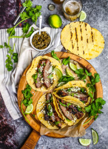 Beautifully plated carne asada tacos with charred beef, fresh cilantro, diced onions, and lime wedges on rustic wooden serving board