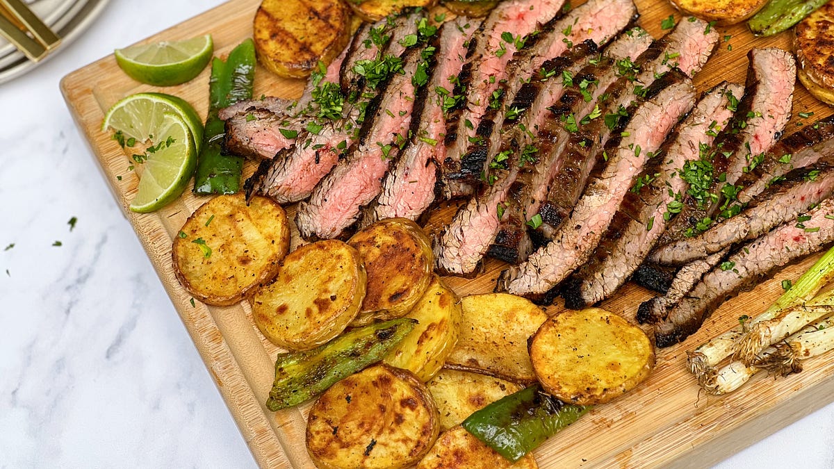 Raw marinated flank steak on cutting board with lime halves, garlic cloves, and fresh cilantro arranged nearby