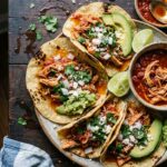 Chicken tinga served in warm corn tortillas topped with fresh cilantro, diced onions, and crumbled queso fresco on a rustic wooden board