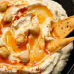Creamy homemade hummus in a white bowl with olive oil drizzled on top, garnished with chickpeas and paprika, surrounded by fresh vegetables and pita bread