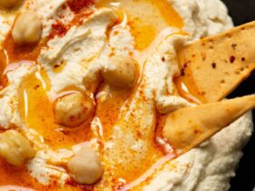 Creamy homemade hummus in a white bowl with olive oil drizzled on top, garnished with chickpeas and paprika, surrounded by fresh vegetables and pita bread