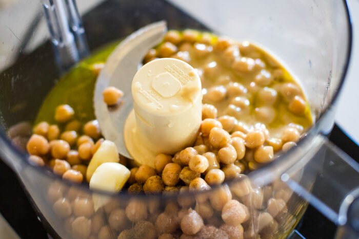 Food processor bowl showing chickpeas being processed into smooth hummus, with tahini and other ingredients visible nearby