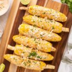 Perfectly grilled Mexican street corn on rustic wooden board with lime wedges and chili powder, showing beautiful char marks and creamy white sauce drizzled over golden kernels