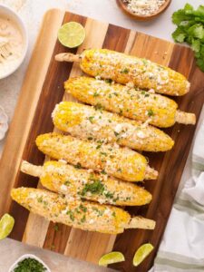 Perfectly grilled Mexican street corn on rustic wooden board with lime wedges and chili powder, showing beautiful char marks and creamy white sauce drizzled over golden kernels