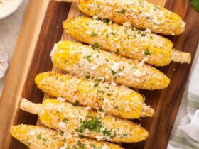 Perfectly grilled Mexican street corn on rustic wooden board with lime wedges and chili powder, showing beautiful char marks and creamy white sauce drizzled over golden kernels