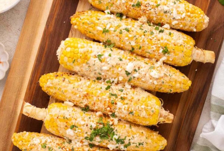 Perfectly grilled Mexican street corn on rustic wooden board with lime wedges and chili powder, showing beautiful char marks and creamy white sauce drizzled over golden kernels