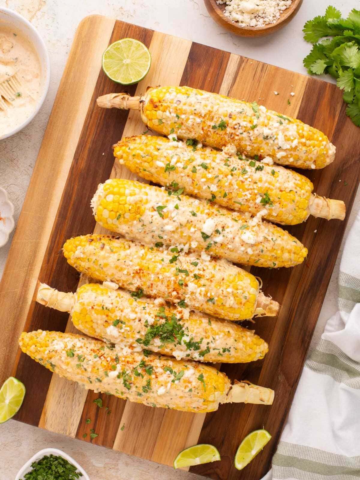 Perfectly grilled Mexican street corn on rustic wooden board with lime wedges and chili powder, showing beautiful char marks and creamy white sauce drizzled over golden kernels