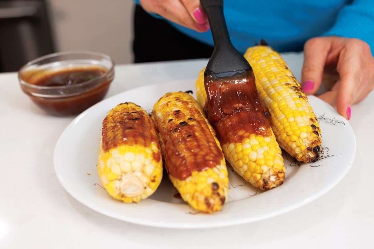 Step-by-step shot showing corn being grilled on barbecue grates with visible char marks forming, alongside a bowl of the yogurt-based crema mixture
