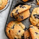 Beautiful golden-brown blueberry muffins with towering tops in a muffin tin, some cooling on a wire rack, with fresh blueberries scattered around