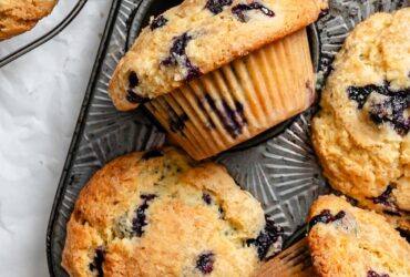 Perfect Blueberry Muffins Recipe - Bakery-Style Fluffy & Bursting with Berries 26 Beautiful golden-brown blueberry muffins with towering tops in a muffin tin, some cooling on a wire rack, with fresh blueberries scattered around