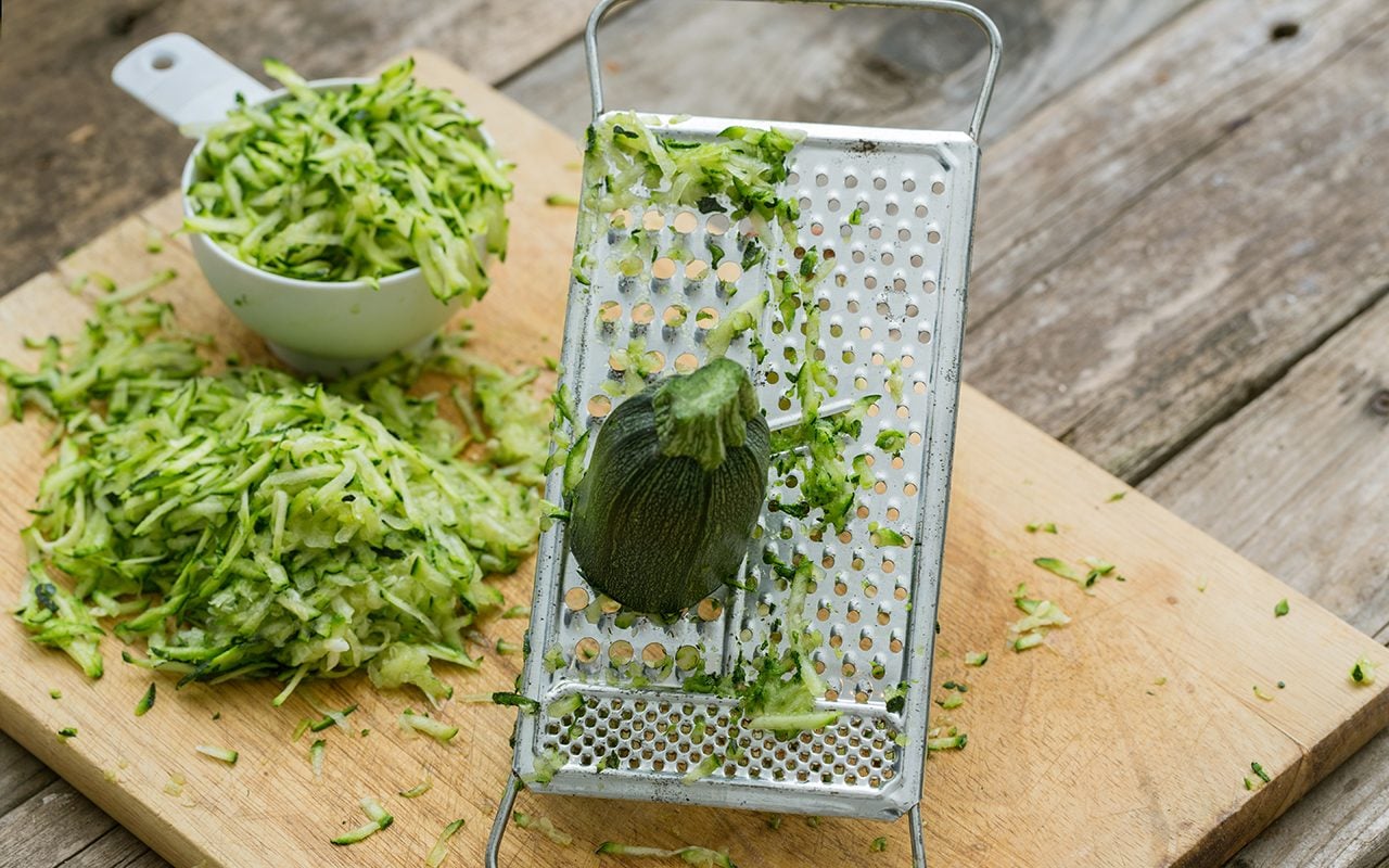 hands grating fresh zucchini on box grater with pile of shredded zucchini