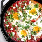 Vibrant shakshuka in cast iron skillet with perfectly poached eggs, fresh herbs, and warm crusty bread on rustic wooden table