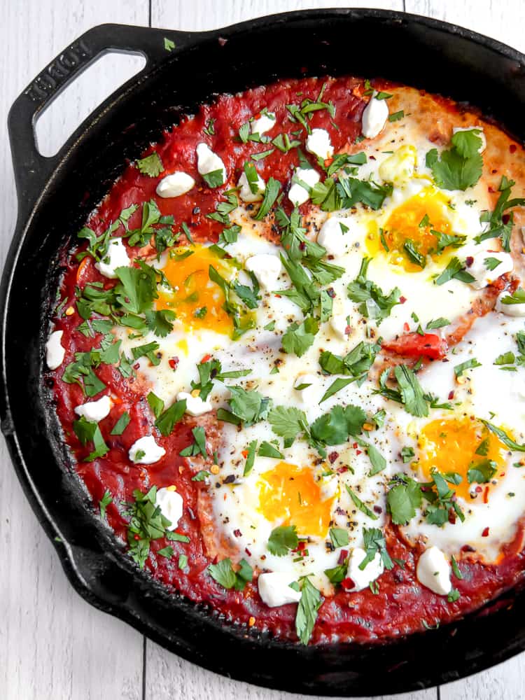 Vibrant shakshuka in cast iron skillet with perfectly poached eggs, fresh herbs, and warm crusty bread on rustic wooden table