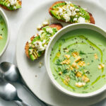 Elegant bowl of pale green cold cucumber avocado soup garnished with fresh herbs, cucumber slices, and a drizzle of olive oil, served on a light wooden board with crusty bread