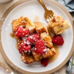 Golden French toast casserole in white baking dish with fresh berries and powdered sugar dusting, styled on rustic wooden table with morning sunlight