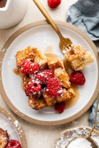 Golden French toast casserole in white baking dish with fresh berries and powdered sugar dusting, styled on rustic wooden table with morning sunlight