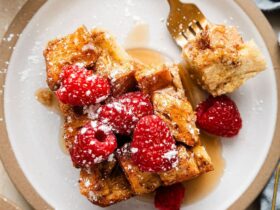 Golden French toast casserole in white baking dish with fresh berries and powdered sugar dusting, styled on rustic wooden table with morning sunlight