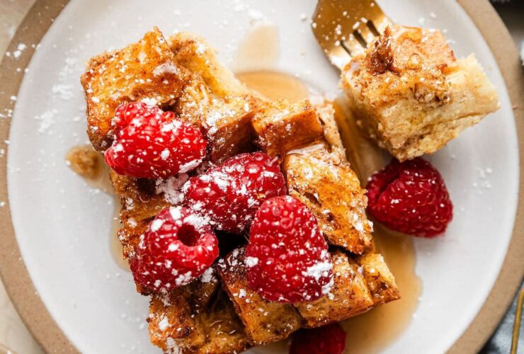Golden French toast casserole in white baking dish with fresh berries and powdered sugar dusting, styled on rustic wooden table with morning sunlight