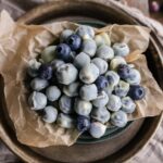 A beautiful overhead flat lay of frozen yogurt covered blueberries arranged on parchment paper with scattered fresh blueberries and a sprig of mint, shot in natural light