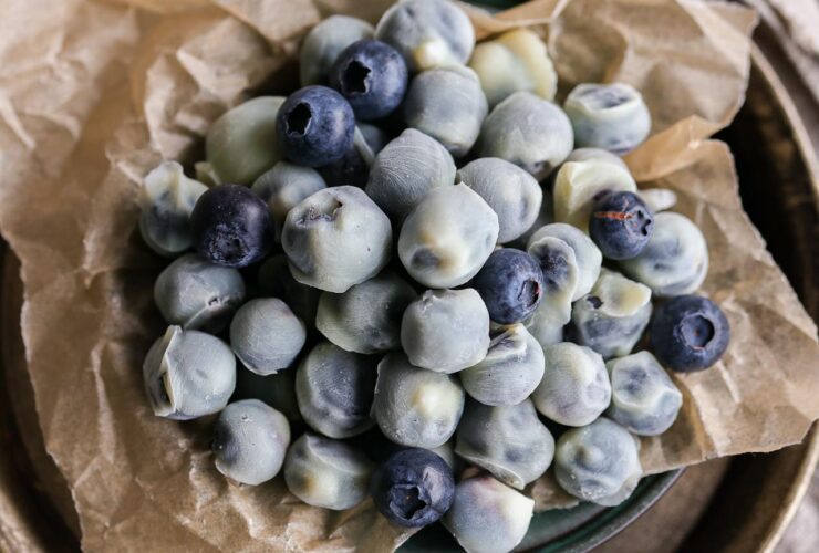A beautiful overhead flat lay of frozen yogurt covered blueberries arranged on parchment paper with scattered fresh blueberries and a sprig of mint, shot in natural light