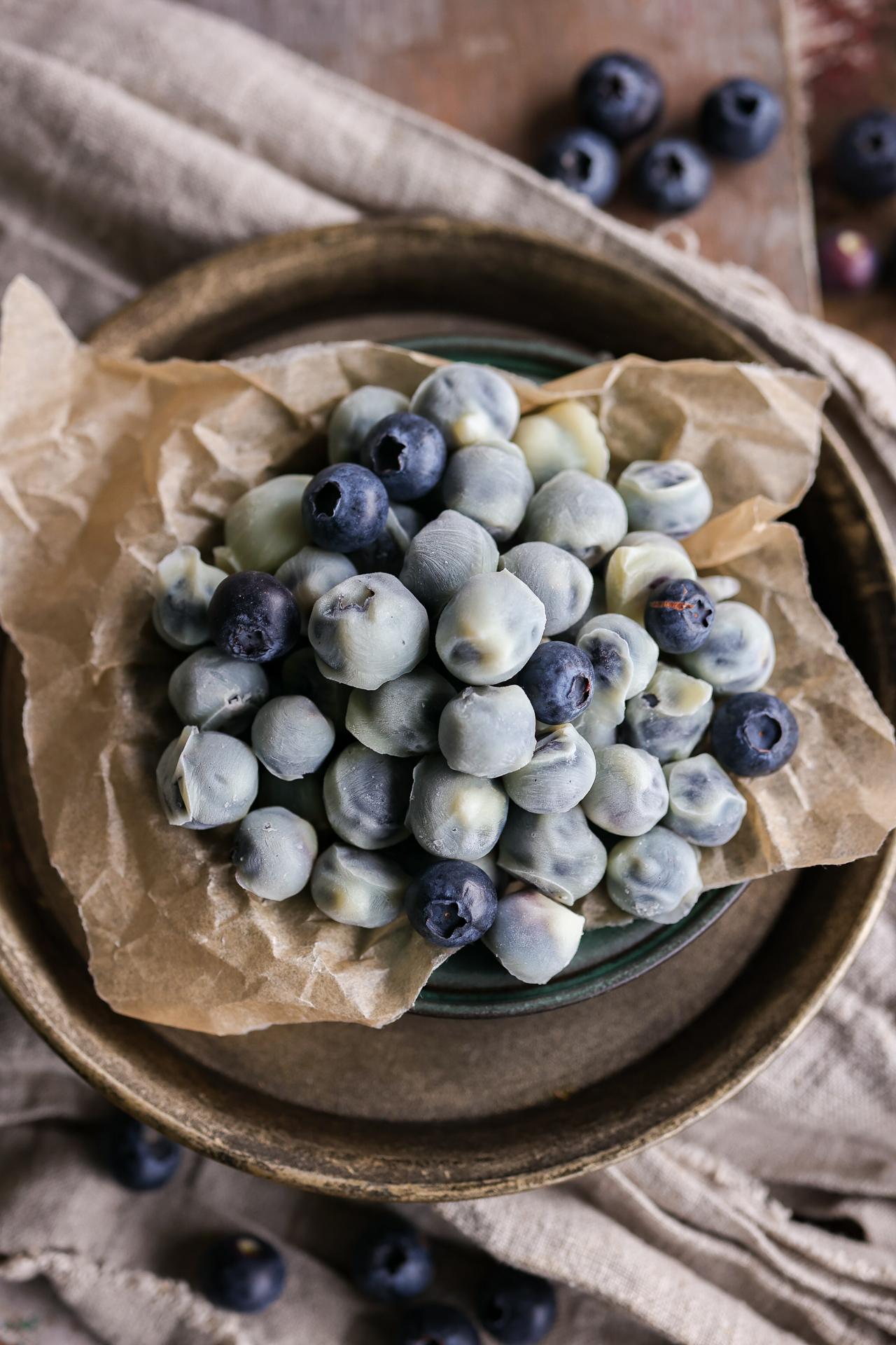 A beautiful overhead flat lay of frozen yogurt covered blueberries arranged on parchment paper with scattered fresh blueberries and a sprig of mint, shot in natural light