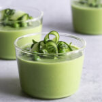Bowl of green gazpacho with avocado garnished with cucumber slices, fresh herbs, a drizzle of olive oil, and microgreens on a white linen background in bright natural light