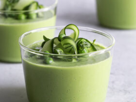 Bowl of green gazpacho with avocado garnished with cucumber slices, fresh herbs, a drizzle of olive oil, and microgreens on a white linen background in bright natural light
