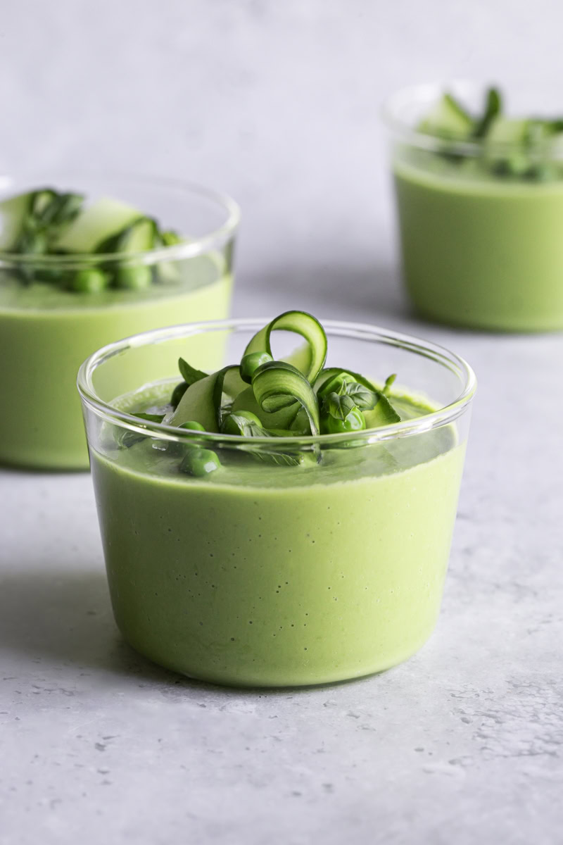 Bowl of green gazpacho with avocado garnished with cucumber slices, fresh herbs, a drizzle of olive oil, and microgreens on a white linen background in bright natural light