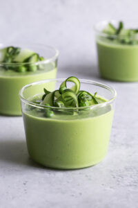 Bowl of green gazpacho with avocado garnished with cucumber slices, fresh herbs, a drizzle of olive oil, and microgreens on a white linen background in bright natural light