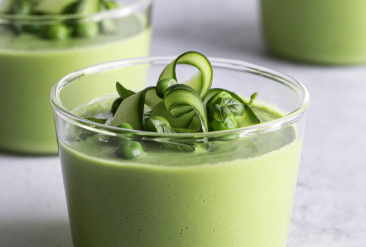 Bowl of green gazpacho with avocado garnished with cucumber slices, fresh herbs, a drizzle of olive oil, and microgreens on a white linen background in bright natural light