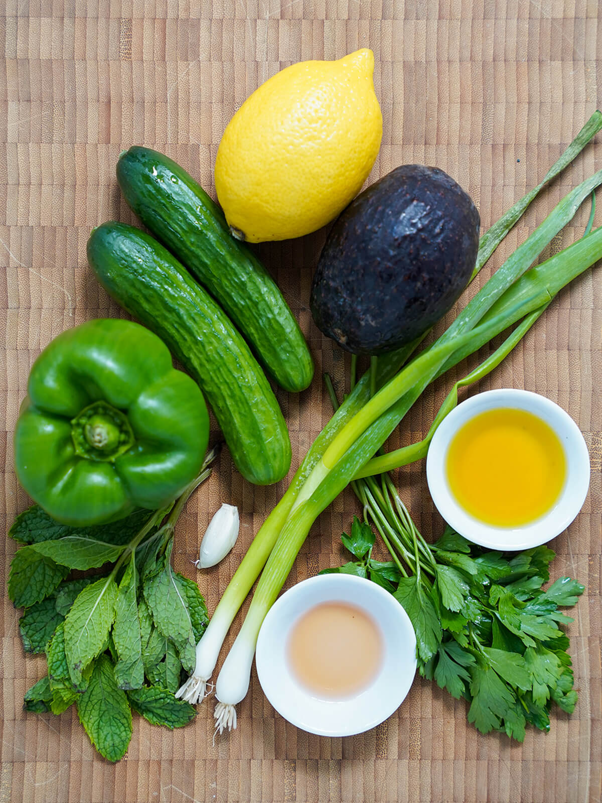 Overhead flat lay of green gazpacho ingredients including halved cucumber, avocado, green bell pepper, garlic cloves, fresh herbs, sherry vinegar bottle, and olive oil on a light marble surface