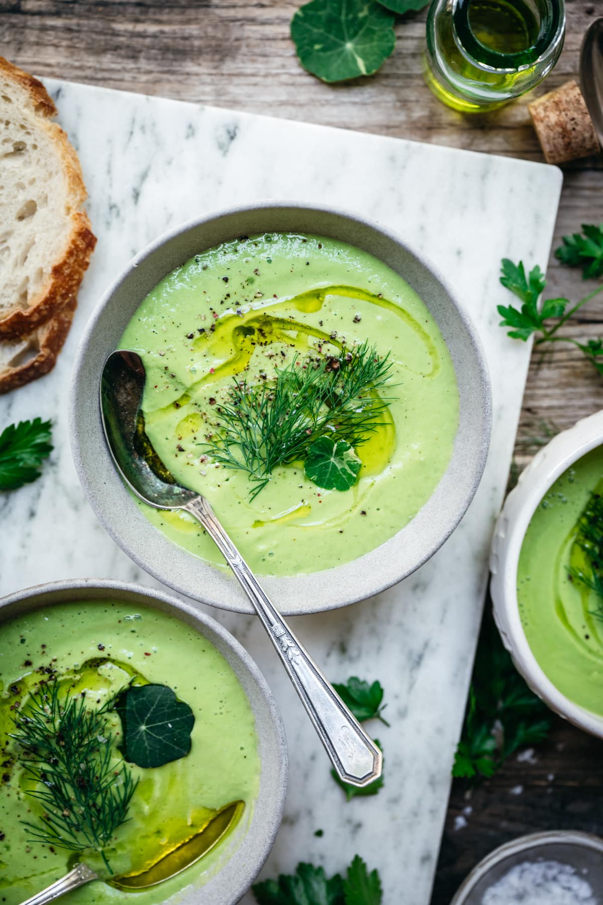 Close-up overhead shot of a white ceramic bowl of green gazpacho with avocado, garnished with thin cucumber ribbons, microgreens, a drizzle of olive oil, and edible flowers on a rustic wooden table