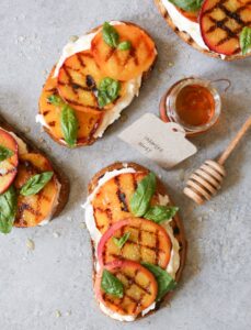 Grilled peach and burrata toast on whole grain bread beautifully plated on rustic wooden board with fresh basil garnish and drizzle of honey