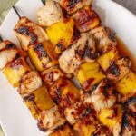 Grilled pineapple chicken skewers beautifully plated on a wooden board with fresh pineapple slices, charred bell peppers, and a sprinkle of fresh cilantro, golden grill marks visible on the chicken