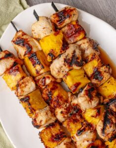 Grilled pineapple chicken skewers beautifully plated on a wooden board with fresh pineapple slices, charred bell peppers, and a sprinkle of fresh cilantro, golden grill marks visible on the chicken