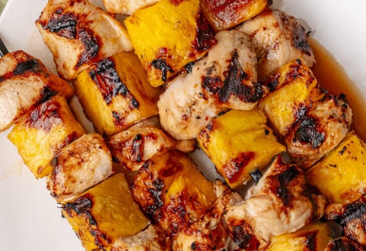 Grilled pineapple chicken skewers beautifully plated on a wooden board with fresh pineapple slices, charred bell peppers, and a sprinkle of fresh cilantro, golden grill marks visible on the chicken