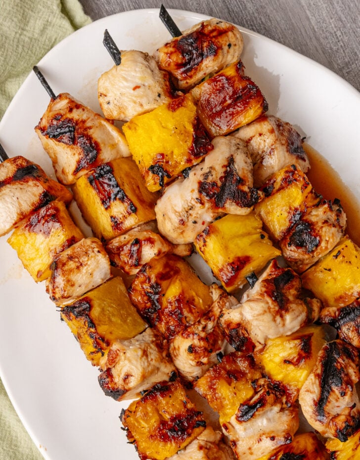 Grilled pineapple chicken skewers beautifully plated on a wooden board with fresh pineapple slices, charred bell peppers, and a sprinkle of fresh cilantro, golden grill marks visible on the chicken