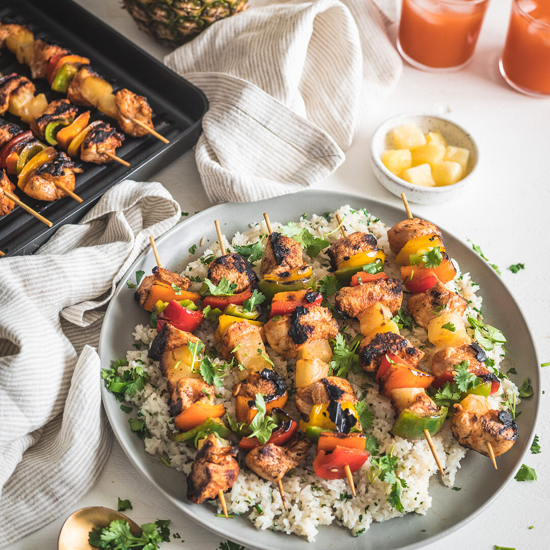 Close-up overhead shot of grilled pineapple chicken skewers served on a platter with a side of cilantro-lime rice, lime wedges, and scattered fresh herbs, warm golden tones with soft natural light