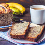 Sliced healthy banana bread on a wooden cutting board with fresh banana slices and a cup of coffee, showing the moist golden interior