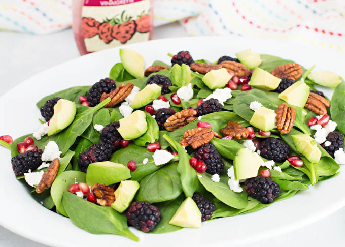Ingredients for healthy summer salad arranged on marble countertop including fresh blackberries, baby spinach, avocado, and small bowl of blackberry balsamic dressing