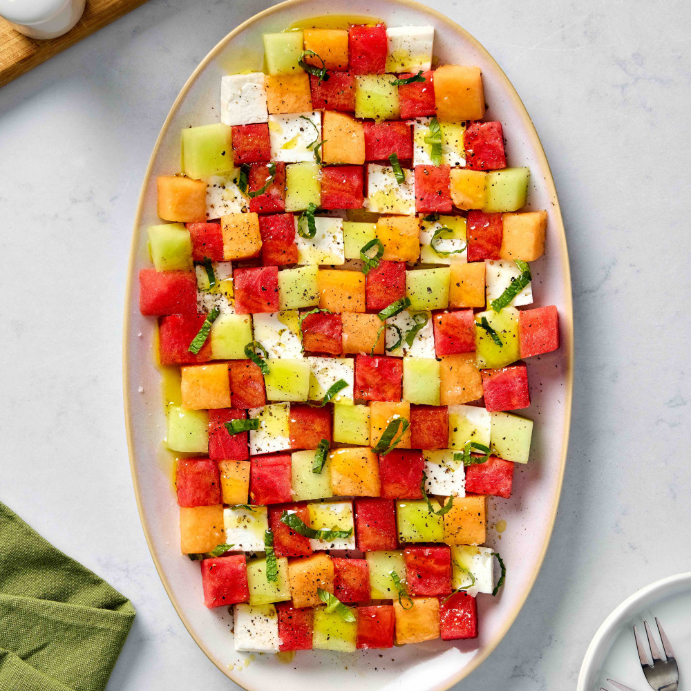 Hands cutting fresh watermelon into cubes on wooden cutting board with mint leaves and feta cheese nearby