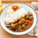 Japanese beef curry served in a white bowl with steamed rice, tender chunks of beef and vegetables visible in rich, golden-brown curry sauce, garnished with fresh herbs