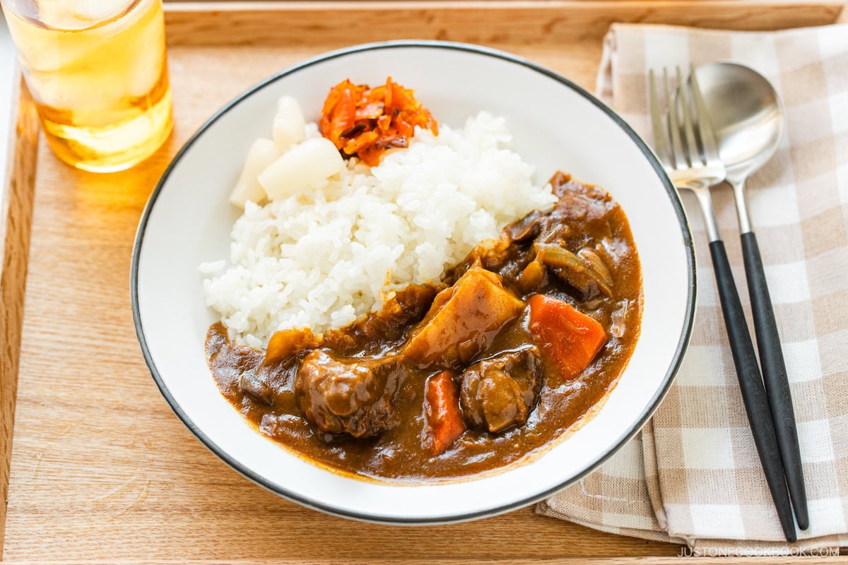 Japanese beef curry served in a white bowl with steamed rice, tender chunks of beef and vegetables visible in rich, golden-brown curry sauce, garnished with fresh herbs