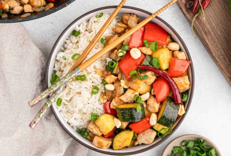 Kung Pao Chicken served in a white bowl with chopsticks, garnished with green onions and crushed peanuts, showing colorful vegetables and tender chicken pieces